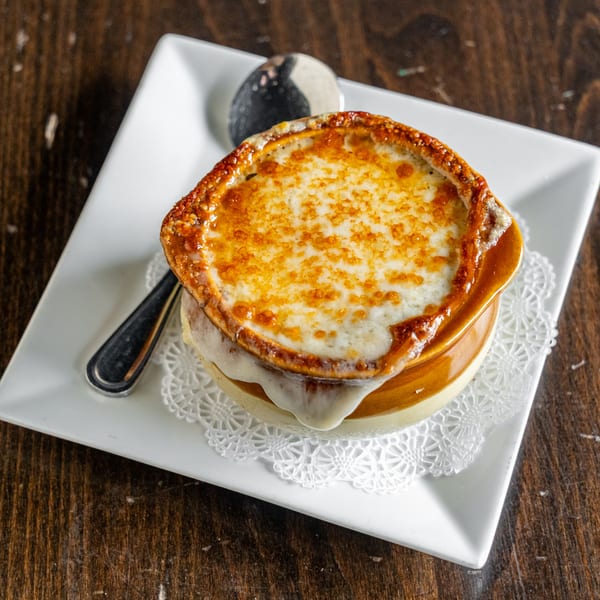 A crock of French onion soup with melted, bubbly cheese on a white plate with a spoon.