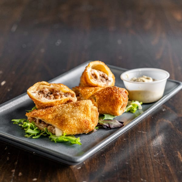 Crispy golden-brown cheesesteak egg rolls with a creamy dipping sauce on a dark plate.