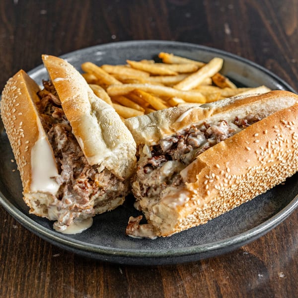 A cheesesteak sandwich, cut in half, with melted cheese and fries on a dark plate.