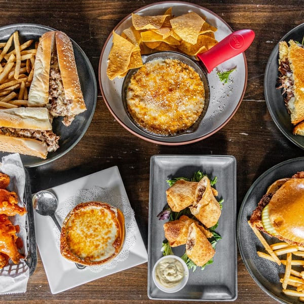 Overhead view of various Steam Pub dishes, including cheesesteaks, dip, egg rolls, and French onion soup.