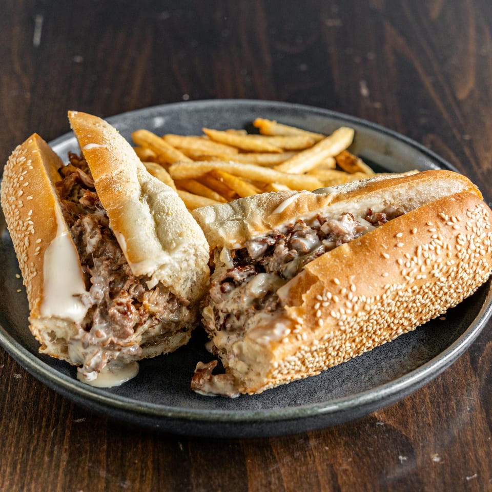 Cheesesteak sandwich cut in half, filled with meat and cheese, served with golden fries on a dark plate.